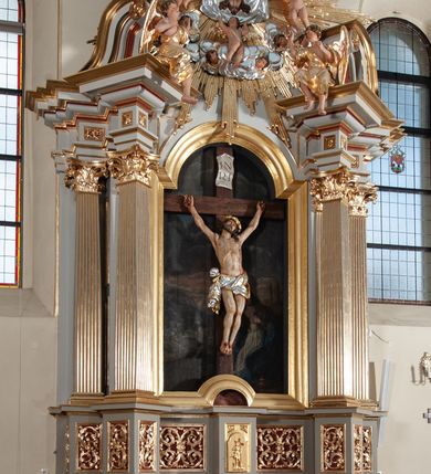 Zdjęcie nr 5: Nastawa ołtarzowa jednoosiowa, jednokondygnacyjna ze zwieńczeniem, na podwójnym cokole. Mensa ołtarzowa drewniana, prostopadłościenna. Antepedium w formie leżącego prostokąta w profilowanej ramie. Pole dekorowane rzeźbionym akantem o układzie symetrycznym względem osi środkowej, z elementami muszli oraz parą puttów po dwóch stronach centralnie ustawionej łodygi z kwiatem. Tabernakulum w formie prostopadłościanu zamkniętego gzymsem. Drzwiczki w kształcie stojącego prostokąta zamkniętego łukiem nadwieszonym w profilowanej ramie z płaskorzeźbionym przedstawieniem zmartwychwstałego Chrystusa; ujęte płyciną, dekorowaną w przyłuczach polami z akantem. Chrystus został ukazany w całej postaci, lewą ręka przytrzymując krzyż, prawą wskazuje ranę w boku. Przy jego prawej nodze stoi kielich. Pierwsza kondygnacja nastawy ujęta dwiema parami kolumn o kapitelach kompozytowych i kanelowanych trzonach o kwadratowym przekroju, ustawionych przed pilastrami; wewnętrzna para ustawiona skośnie względem lica ołtarza. Podpory dźwigają pełne belkowanie wyłamane na ich osi, przerwane nad polem środkowym; wewnętrzna para podtrzymuje imposty oraz przerwany, esowaty przyczółek z siedzącymi przed nim, na gzymsie, figurami aniołów w długich sukniach. Pole środkowe kondygnacji w formie stojącego prostokąta zamkniętego łukiem półkolistym nadwieszonym z wnęką z figurą Chrystusa Ukrzyżowanego na malowanym tle z Jerozolimą i postacią św. Marii Magdaleny. Zwieńczenie w formie bardzo rozbudowanej glorii promienistej, o naprzemiennie długich i krótkich wiązkach promieni; z płaskorzeźbionym mandylionem podtrzymywanym u spodu i po bokach przez trzy putta. Chustę św. Weroniki od góry i od dołu ujmują skłębione obłoki, między którymi widoczne główki anielskie. 
Oba cokoły dekorowane płycinami: w partii dolnego cokołu wypełnionymi ciemnoróżową marmoryzacją, w partii górnego ornamentem akantowym, mniejsze płyciny w partii fryzu. Struktura polichromowana na kolor jasnoszary, pola płycin i profilowania czerwone; kolumny, gloria, ornamentyka i profilowania złocone; chusta i obłoki srebrzone.
