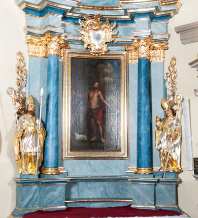 Zdjęcie nr 1: Nastawa ołtarzowa jednoosiowa, jednokondygnacyjna ze zwieńczeniem, na podwójnym cokole. Mensa drewniana, prostopadłościenna. Antepedium w formie leżącego prostokąta ujęte profilowaną ramą. W polu płaskorzeźbiona półpostać św. Augustyna ujętego u spodu płatkami kwiatu, flankowana banderolami. W prawej, uniesionej ręce trzyma księgę, lewą, ugiętą przytrzymuje pastorał, dłonią przyciska do piersi gorejące serce. Święty ukazany został jako starzec. Twarz ma pociągłą, okoloną krótką brodą, włosy sięgające szyi. Ubrany jest w niebieską tunikę i przerzucony przez ramiona płaszcz ze złoconą lamówką. Pole wypełnione jest płaskorzeźbionymi łodygami z kwiatami. Kondygnacja nastawy o wklęsłym licu, ujęta parą zwielokrotnionych pilastrów oraz stojącą przed nimi parą kolumn o kapitelach kompozytowych. Podpory dźwigają pełne belkowanie o silnie wysuniętym gzymsie, wyłamane na ich osi oraz przerwany przyczółek na linii kolumn. Po zewnętrznej stronie kolumn figury św. Wojciecha po lewej i św. Stanisława po prawej. W polu środkowym nisza w formie stojącego prostokąta otoczona profilowaną ramą; wewnątrz mniejszy obraz z przedstawieniem św. Jana Chrzciciela w profilowanym obramieniu w kształcie stojącego prostokąta. Zwieńczenie w kształcie stojącego prostokąta ujętego spływami wolutowymi, zamknięte gzymsem utworzonym ze zwróconych do siebie wolut; zakończone glorią promienistą z okrągłym polem z napisem „IHS” w otoczeniu obłoków. W polu zwieńczenia obraz z przedstawieniem św. Grzegorza w profilowanej ramie w formie stojącego prostokąta o wklęsłych bokach i narożach, zamkniętej łukiem nadwieszonym. Kondygnacja ujęta ażurowymi uszami utworzonymi z motywów roślinnych i karbowanej wstęgi. W partii belkowania nad polem środkowym rocaille'owy kartusz. Nad obrazem w zwieńczeniu muszla i kampanule; woluty dekorowane festonami. Struktura polichromowana na kolor jasnoniebieski; trzony kolumn, antepedium i profile na niebiesko; ornamentyka i profile złocone.