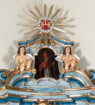 Zdjęcie nr 6: Nastawa ołtarzowa jednoosiowa, jednokondygnacyjna ze zwieńczeniem, na podwójnym cokole. Mensa drewniana, prostopadłościenna. Antepedium w formie leżącego prostokąta ujęte profilowaną ramą. W polu płaskorzeźbiona półpostać św. Augustyna ujętego u spodu płatkami kwiatu, flankowana banderolami. W prawej, uniesionej ręce trzyma księgę, lewą, ugiętą przytrzymuje pastorał, dłonią przyciska do piersi gorejące serce. Święty ukazany został jako starzec. Twarz ma pociągłą, okoloną krótką brodą, włosy sięgające szyi. Ubrany jest w niebieską tunikę i przerzucony przez ramiona płaszcz ze złoconą lamówką. Pole wypełnione jest płaskorzeźbionymi łodygami z kwiatami. Kondygnacja nastawy o wklęsłym licu, ujęta parą zwielokrotnionych pilastrów oraz stojącą przed nimi parą kolumn o kapitelach kompozytowych. Podpory dźwigają pełne belkowanie o silnie wysuniętym gzymsie, wyłamane na ich osi oraz przerwany przyczółek na linii kolumn. Po zewnętrznej stronie kolumn figury św. Wojciecha po lewej i św. Stanisława po prawej. W polu środkowym nisza w formie stojącego prostokąta otoczona profilowaną ramą; wewnątrz mniejszy obraz z przedstawieniem św. Jana Chrzciciela w profilowanym obramieniu w kształcie stojącego prostokąta. Zwieńczenie w kształcie stojącego prostokąta ujętego spływami wolutowymi, zamknięte gzymsem utworzonym ze zwróconych do siebie wolut; zakończone glorią promienistą z okrągłym polem z napisem „IHS” w otoczeniu obłoków. W polu zwieńczenia obraz z przedstawieniem św. Grzegorza w profilowanej ramie w formie stojącego prostokąta o wklęsłych bokach i narożach, zamkniętej łukiem nadwieszonym. Kondygnacja ujęta ażurowymi uszami utworzonymi z motywów roślinnych i karbowanej wstęgi. W partii belkowania nad polem środkowym rocaille&#039;owy kartusz. Nad obrazem w zwieńczeniu muszla i kampanule; woluty dekorowane festonami. Struktura polichromowana na kolor jasnoniebieski; trzony kolumn, antepedium i profile na niebiesko; ornamentyka i profile złocone.