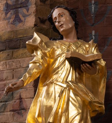 Zdjęcie nr 1: Rzeźba drążona na cokole z przedstawieniem św. Jana Ewangelisty w całej postaci. Figura w kontrapoście, w skręcie w prawo, z głową zwróconą w tę samą stronę; w lewej wyciągniętej w bok ręce trzyma księgę, prawą unosi do boku. Głowę ma lekko przechyloną; twarz młodzieńczą, szczupłą z wysokim czołem, o szeroko otwartych, ciemnych oczach, wąskim nosie i niewielkich ustach. Twarz jest okolona włosami układającymi się w spływające na plecy fale. Święty jest ubrany w długą, przylegającą do ciała tunikę z kołnierzykiem oraz płaszcz przewiązany diagonalnie z prawego ramienia przez plecy i wokół lewego biodra, zatknięty z przodu za pasek. Polichromia naturalistyczna w odsłoniętych partiach ciała, szaty złocone, księga brązowo-złota, karty białe. Z uwagi na swoje aktualne ustawienie na ołtarzu głównym wydrążenie zostało zasłonięte zamalowanym na złoto materiałem.