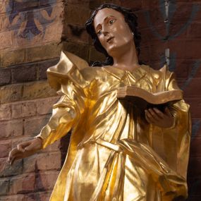 Zdjęcie nr 1: Rzeźba drążona na cokole z przedstawieniem św. Jana Ewangelisty w całej postaci. Figura w kontrapoście, w skręcie w prawo, z głową zwróconą w tę samą stronę; w lewej wyciągniętej w bok ręce trzyma księgę, prawą unosi do boku. Głowę ma lekko przechyloną; twarz młodzieńczą, szczupłą z wysokim czołem, o szeroko otwartych, ciemnych oczach, wąskim nosie i niewielkich ustach. Twarz jest okolona włosami układającymi się w spływające na plecy fale. Święty jest ubrany w długą, przylegającą do ciała tunikę z kołnierzykiem oraz płaszcz przewiązany diagonalnie z prawego ramienia przez plecy i wokół lewego biodra, zatknięty z przodu za pasek. Polichromia naturalistyczna w odsłoniętych partiach ciała, szaty złocone, księga brązowo-złota, karty białe. Z uwagi na swoje aktualne ustawienie na ołtarzu głównym wydrążenie zostało zasłonięte zamalowanym na złoto materiałem.