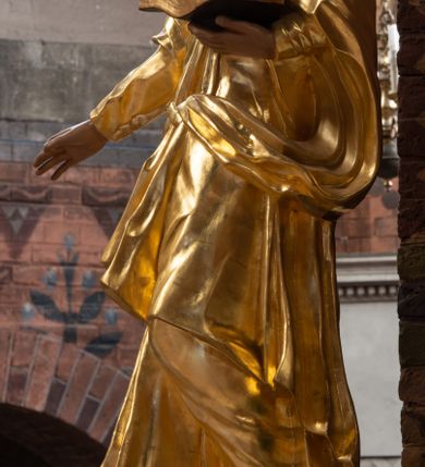 Zdjęcie nr 2: Rzeźba drążona na cokole z przedstawieniem św. Jana Ewangelisty w całej postaci. Figura w kontrapoście, w skręcie w prawo, z głową zwróconą w tę samą stronę; w lewej wyciągniętej w bok ręce trzyma księgę, prawą unosi do boku. Głowę ma lekko przechyloną; twarz młodzieńczą, szczupłą z wysokim czołem, o szeroko otwartych, ciemnych oczach, wąskim nosie i niewielkich ustach. Twarz jest okolona włosami układającymi się w spływające na plecy fale. Święty jest ubrany w długą, przylegającą do ciała tunikę z kołnierzykiem oraz płaszcz przewiązany diagonalnie z prawego ramienia przez plecy i wokół lewego biodra, zatknięty z przodu za pasek. Polichromia naturalistyczna w odsłoniętych partiach ciała, szaty złocone, księga brązowo-złota, karty białe. Z uwagi na swoje aktualne ustawienie na ołtarzu głównym wydrążenie zostało zasłonięte zamalowanym na złoto materiałem.