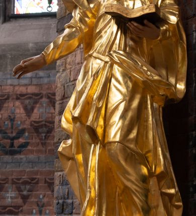 Zdjęcie nr 6: Rzeźba drążona na cokole z przedstawieniem św. Jana Ewangelisty w całej postaci. Figura w kontrapoście, w skręcie w prawo, z głową zwróconą w tę samą stronę; w lewej wyciągniętej w bok ręce trzyma księgę, prawą unosi do boku. Głowę ma lekko przechyloną; twarz młodzieńczą, szczupłą z wysokim czołem, o szeroko otwartych, ciemnych oczach, wąskim nosie i niewielkich ustach. Twarz jest okolona włosami układającymi się w spływające na plecy fale. Święty jest ubrany w długą, przylegającą do ciała tunikę z kołnierzykiem oraz płaszcz przewiązany diagonalnie z prawego ramienia przez plecy i wokół lewego biodra, zatknięty z przodu za pasek. Polichromia naturalistyczna w odsłoniętych partiach ciała, szaty złocone, księga brązowo-złota, karty białe. Z uwagi na swoje aktualne ustawienie na ołtarzu głównym wydrążenie zostało zasłonięte zamalowanym na złoto materiałem.