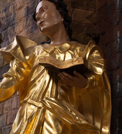 Zdjęcie nr 4: Rzeźba drążona na cokole z przedstawieniem św. Jana Ewangelisty w całej postaci. Figura w kontrapoście, w skręcie w prawo, z głową zwróconą w tę samą stronę; w lewej wyciągniętej w bok ręce trzyma księgę, prawą unosi do boku. Głowę ma lekko przechyloną; twarz młodzieńczą, szczupłą z wysokim czołem, o szeroko otwartych, ciemnych oczach, wąskim nosie i niewielkich ustach. Twarz jest okolona włosami układającymi się w spływające na plecy fale. Święty jest ubrany w długą, przylegającą do ciała tunikę z kołnierzykiem oraz płaszcz przewiązany diagonalnie z prawego ramienia przez plecy i wokół lewego biodra, zatknięty z przodu za pasek. Polichromia naturalistyczna w odsłoniętych partiach ciała, szaty złocone, księga brązowo-złota, karty białe. Z uwagi na swoje aktualne ustawienie na ołtarzu głównym wydrążenie zostało zasłonięte zamalowanym na złoto materiałem.