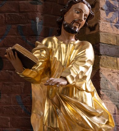 Zdjęcie nr 2: Rzeźba drążona na cokole z przedstawieniem św. Łukasza w całej postaci. Figura w kontrapoście, w skręcie w prawo, z głową zwróconą w lewo; w prawej wyciągniętej w bok ręce trzyma księgę, lewą unosi równolegle. Głowę ma lekko pochyloną; twarz szczupłą z wysokim czołem, o szeroko otwartych, ciemnych oczach, wąskim nosie i rozchylonych ustach. Twarz jest okolona krótkim kręconym zarostem oraz włosami układającymi się w spływające na kark fale. Święty jest ubrany w długą, przylegającą do ciała tunikę oraz płaszcz przewiązany diagonalnie z prawego ramienia przez plecy i wokół lewego biodra, złapany z przodu. Polichromia naturalistyczna w odsłoniętych partiach ciała, szaty złocone, księga brązowo-złota. Z uwagi na swoje aktualne ustawienie na ołtarzu głównym wydrążenie zostało zasłonięte zamalowanym na złoto materiałem.