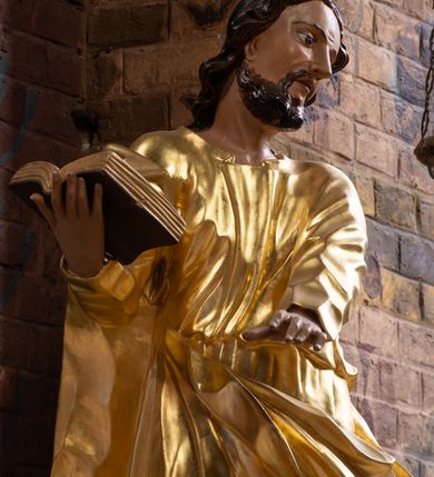 Zdjęcie nr 3: Rzeźba drążona na cokole z przedstawieniem św. Łukasza w całej postaci. Figura w kontrapoście, w skręcie w prawo, z głową zwróconą w lewo; w prawej wyciągniętej w bok ręce trzyma księgę, lewą unosi równolegle. Głowę ma lekko pochyloną; twarz szczupłą z wysokim czołem, o szeroko otwartych, ciemnych oczach, wąskim nosie i rozchylonych ustach. Twarz jest okolona krótkim kręconym zarostem oraz włosami układającymi się w spływające na kark fale. Święty jest ubrany w długą, przylegającą do ciała tunikę oraz płaszcz przewiązany diagonalnie z prawego ramienia przez plecy i wokół lewego biodra, złapany z przodu. Polichromia naturalistyczna w odsłoniętych partiach ciała, szaty złocone, księga brązowo-złota. Z uwagi na swoje aktualne ustawienie na ołtarzu głównym wydrążenie zostało zasłonięte zamalowanym na złoto materiałem.