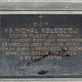 Zdjęcie nr 1: Kamienna tablica w kształcie leżącego prostokąta w szerokiej, profilowanej ramie z uszakami. Na niej, poniżej niewielkiego krzyża łacińskiego majuskułowy, złocony napis „D(EO) O(PTIMO) M(AXIMO) / KS(IĄDZ) MICHAŁ KOŁODZIEJ / DOKTÓR FILOZOFJI I PRAWA / BYŁY KANONIK KAPITUŁY KRAKOWSKIEJ / SZAMBELAN JEGO ŚWIĄTOBLIWOŚCI / DZIEKAN SUSKI I PROBOSZCZ W SUCHEJ / ZMARŁ 25 XI 1928 W 58 ROKU ŻYCIA / A 28 KAPŁAŃSTWA. / CZEŚĆ JEGO PAMIĘCI”.