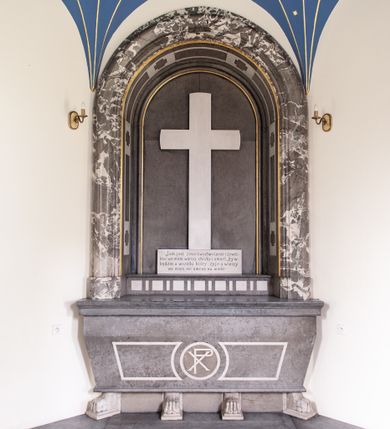 Zdjęcie nr 1: Mensa w typie sarkofagowym ustawiona na czterech lwich łapach. Ściana frontowa jest dekorowana trzema płycinami. Zewnętrzne mają kształt leżących prostokątów z wklęsłą wewnętrzną krawędzią, a środkowa jest okrągła, wypełniona chrystogramem. Mensa w kolorze jasnoszarego kamienia, łapy białe. 