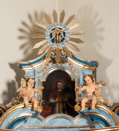 Zdjęcie nr 2: Obraz w kształcie stojącego prostokąta o wklęsłych bokach i dolnych narożach, zamknięty łukiem nadwieszonym, w profilowanej, złoconej i srebrzonej ramie. W centrum kompozycji stoi św. Grzegorz, ukazany do bioder, zwrócony w trzech czwartych w lewo, z prawą ręką uniesioną w geście błogosławieństwa, w lewej trzyma krzyż potrójny pontyfikalny. Ma owalną twarz z wyraźnymi bruzdami zmarszczek. Przylegające nakrycie, które ma na głowie nosi nazwę camerino (camauro), na obrazie widać, że jest ubrany w rokietę, na którą ma założony mucet – sięgającą łokci pelerynę i złotą stułę. Na drugim planie, na stole po lewej stronie stoi niebiesko-złota tiara papieska. Tło obrazu ciemnobrunatne, rozjaśnione subtelnie za głową postaci.  