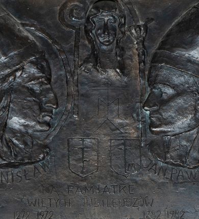 Zdjęcie nr 1: Brązowa tablica w kształcie leżącego prostokątna obwiedzioną szeroką, profilowaną, kamienną ramą. Na niej symetrycznie po dwóch stronach profilowe portrety zwróconych do siebie: po lewej stronie św. Stanisława, a po prawej św. Jana Pawła. Święty Stanisław ukazany w nimbie i z pastorałem, św. Jan Paweł II z krzyżem papieskim. Obaj zostali przedstawieni w infułach i podpisani „ŚW(IĘTY) STANISŁAW” i „ JAN PAWEŁ II P(APIEŻ)P(OLAK)”. Między nimi, u góry, niewielkie portretowe ujęcie Matki Boskiej Częstochowskiej, poniżej której jej monogram ponad herbami wymienionych postaci. Na dole, w trzech linijkach napis „NA PAMIATKĘ / ŚWIĘTYCH JUBILEUSZÓW / 1272 1972 1983  1382 1982”. W prawym dolnym rogu sygnatura „B. LANGMAN”. 