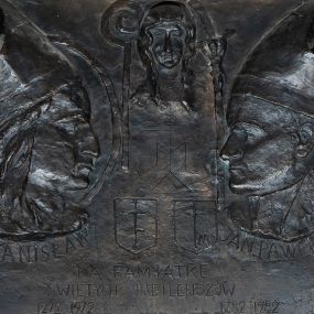 Zdjęcie nr 1: Brązowa tablica w kształcie leżącego prostokątna obwiedzioną szeroką, profilowaną, kamienną ramą. Na niej symetrycznie po dwóch stronach profilowe portrety zwróconych do siebie: po lewej stronie św. Stanisława, a po prawej św. Jana Pawła. Święty Stanisław ukazany w nimbie i z pastorałem, św. Jan Paweł II z krzyżem papieskim. Obaj zostali przedstawieni w infułach i podpisani „ŚW(IĘTY) STANISŁAW” i „ JAN PAWEŁ II P(APIEŻ)P(OLAK)”. Między nimi, u góry, niewielkie portretowe ujęcie Matki Boskiej Częstochowskiej, poniżej której jej monogram ponad herbami wymienionych postaci. Na dole, w trzech linijkach napis „NA PAMIATKĘ / ŚWIĘTYCH JUBILEUSZÓW / 1272 1972 1983  1382 1982”. W prawym dolnym rogu sygnatura „B. LANGMAN”. 