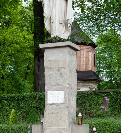 Zdjęcie nr 4: Pełnoplastyczna rzeźba Chrystusa Króla Wszechświata została ustawiona na wysokim czworobocznym postumencie wzniesionym z kamiennych ciosów. Chrystus został przedstawiony frontalnie, stojąc na fragmencie widocznego globu w nieznacznym kontrapoście. W wyciągniętej przed siebie prawej ręce trzyma złocone berło ułożone poziomo. Natomiast lewą ręką podtrzymuje niebieski glob zwieńczony złotym krzyżykiem. Chrystus ma pociągłą twarz otoczoną zarostem oraz długimi włosami spływającymi na ramiona. Ma prosty nos, wydatne usta oraz szeroko otwarte oczy o spojrzeniu skierowanym przed siebie. Ubrany jest w przewiązaną albę odsłaniającą na dole końcówki butów, skrzyżowaną na piersi kapłańską stułę oraz kapę, której poły zarzucone są na przedramienia. Spiętą pod szyją złoconą fibulą kapę lamuje szeroka, wzorzysta borta wyciskana w kamieniu. Zbawiciel ma na głowie złoconą koronę o wysokich, floralnych sterczynach z obręczą dekorowaną motywem wieńca z ciernia. Boki postumentu flankują dwa kamienne uskoki ułożone schodkowo. 
