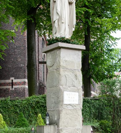 Zdjęcie nr 2: Pełnoplastyczna rzeźba Chrystusa Króla Wszechświata została ustawiona na wysokim czworobocznym postumencie wzniesionym z kamiennych ciosów. Chrystus został przedstawiony frontalnie, stojąc na fragmencie widocznego globu w nieznacznym kontrapoście. W wyciągniętej przed siebie prawej ręce trzyma złocone berło ułożone poziomo. Natomiast lewą ręką podtrzymuje niebieski glob zwieńczony złotym krzyżykiem. Chrystus ma pociągłą twarz otoczoną zarostem oraz długimi włosami spływającymi na ramiona. Ma prosty nos, wydatne usta oraz szeroko otwarte oczy o spojrzeniu skierowanym przed siebie. Ubrany jest w przewiązaną albę odsłaniającą na dole końcówki butów, skrzyżowaną na piersi kapłańską stułę oraz kapę, której poły zarzucone są na przedramienia. Spiętą pod szyją złoconą fibulą kapę lamuje szeroka, wzorzysta borta wyciskana w kamieniu. Zbawiciel ma na głowie złoconą koronę o wysokich, floralnych sterczynach z obręczą dekorowaną motywem wieńca z ciernia. Boki postumentu flankują dwa kamienne uskoki ułożone schodkowo. 