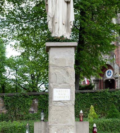 Zdjęcie nr 3: Pełnoplastyczna rzeźba Chrystusa Króla Wszechświata została ustawiona na wysokim czworobocznym postumencie wzniesionym z kamiennych ciosów. Chrystus został przedstawiony frontalnie, stojąc na fragmencie widocznego globu w nieznacznym kontrapoście. W wyciągniętej przed siebie prawej ręce trzyma złocone berło ułożone poziomo. Natomiast lewą ręką podtrzymuje niebieski glob zwieńczony złotym krzyżykiem. Chrystus ma pociągłą twarz otoczoną zarostem oraz długimi włosami spływającymi na ramiona. Ma prosty nos, wydatne usta oraz szeroko otwarte oczy o spojrzeniu skierowanym przed siebie. Ubrany jest w przewiązaną albę odsłaniającą na dole końcówki butów, skrzyżowaną na piersi kapłańską stułę oraz kapę, której poły zarzucone są na przedramienia. Spiętą pod szyją złoconą fibulą kapę lamuje szeroka, wzorzysta borta wyciskana w kamieniu. Zbawiciel ma na głowie złoconą koronę o wysokich, floralnych sterczynach z obręczą dekorowaną motywem wieńca z ciernia. Boki postumentu flankują dwa kamienne uskoki ułożone schodkowo. 