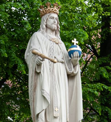 Zdjęcie nr 1: Pełnoplastyczna rzeźba Chrystusa Króla Wszechświata została ustawiona na wysokim czworobocznym postumencie wzniesionym z kamiennych ciosów. Chrystus został przedstawiony frontalnie, stojąc na fragmencie widocznego globu w nieznacznym kontrapoście. W wyciągniętej przed siebie prawej ręce trzyma złocone berło ułożone poziomo. Natomiast lewą ręką podtrzymuje niebieski glob zwieńczony złotym krzyżykiem. Chrystus ma pociągłą twarz otoczoną zarostem oraz długimi włosami spływającymi na ramiona. Ma prosty nos, wydatne usta oraz szeroko otwarte oczy o spojrzeniu skierowanym przed siebie. Ubrany jest w przewiązaną albę odsłaniającą na dole końcówki butów, skrzyżowaną na piersi kapłańską stułę oraz kapę, której poły zarzucone są na przedramienia. Spiętą pod szyją złoconą fibulą kapę lamuje szeroka, wzorzysta borta wyciskana w kamieniu. Zbawiciel ma na głowie złoconą koronę o wysokich, floralnych sterczynach z obręczą dekorowaną motywem wieńca z ciernia. Boki postumentu flankują dwa kamienne uskoki ułożone schodkowo. 