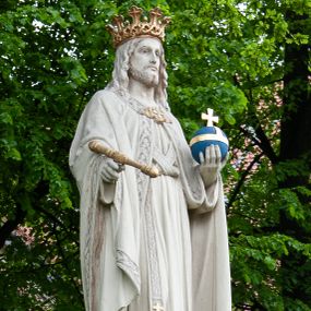 Zdjęcie nr 1: Pełnoplastyczna rzeźba Chrystusa Króla Wszechświata została ustawiona na wysokim czworobocznym postumencie wzniesionym z kamiennych ciosów. Chrystus został przedstawiony frontalnie, stojąc na fragmencie widocznego globu w nieznacznym kontrapoście. W wyciągniętej przed siebie prawej ręce trzyma złocone berło ułożone poziomo. Natomiast lewą ręką podtrzymuje niebieski glob zwieńczony złotym krzyżykiem. Chrystus ma pociągłą twarz otoczoną zarostem oraz długimi włosami spływającymi na ramiona. Ma prosty nos, wydatne usta oraz szeroko otwarte oczy o spojrzeniu skierowanym przed siebie. Ubrany jest w przewiązaną albę odsłaniającą na dole końcówki butów, skrzyżowaną na piersi kapłańską stułę oraz kapę, której poły zarzucone są na przedramienia. Spiętą pod szyją złoconą fibulą kapę lamuje szeroka, wzorzysta borta wyciskana w kamieniu. Zbawiciel ma na głowie złoconą koronę o wysokich, floralnych sterczynach z obręczą dekorowaną motywem wieńca z ciernia. Boki postumentu flankują dwa kamienne uskoki ułożone schodkowo. 