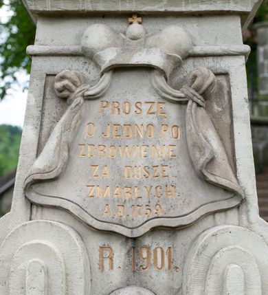 Zdjęcie nr 5: Figura św. Jana Nepomucena na rozbudowanej podstawie. Na trzech stopniach ustawiono sześcian zwężający się ku górze, składający się z postumentu z wolutami w dolnej części ustawionego na cokole i zwieńczonego gzymsem. Na ścianie frontowej na rozpostartej, zwieńczonej mitrą książęcą tkaninie złocona inskrypcja „PROSZĘ / O JEDNO PO/ZDROWIENIE / ZA DUSZE / ZMARŁYCH / A(NNO) D(OMINI) 1759” a poniżej data „R(OKU) 1901” ponad czaszką i piszczelą. Na podstawie, na cokole ustawiono figurę św. Jana Nepomucena w kontrapoście, z prawą nogą ugiętą w kolanie. Obiema rękami przytrzymuje krucyfiks i złotą palmę męczeństwa. Ma podłużną, smutną twarz o wklęsłych policzkach, niewielkich oczach, wąskim nosie i zamkniętych ustach okoloną krótką brodą i krótkimi włosami odwijającymi się do tyłu. Jest ubrany w strój kanonika: ma narzuconą na komżę i sutannę futrzaną almucję, a na głowie biret. Wokół głowy nimb z pięcioma złoconymi gwiazdkami. Na stopniu cokoło napis „ODNOWIŁA RADA GMINNA”. 