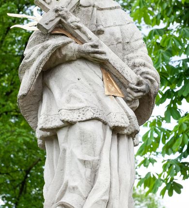 Zdjęcie nr 4: Figura św. Jana Nepomucena na rozbudowanej podstawie. Na trzech stopniach ustawiono sześcian zwężający się ku górze, składający się z postumentu z wolutami w dolnej części ustawionego na cokole i zwieńczonego gzymsem. Na ścianie frontowej na rozpostartej, zwieńczonej mitrą książęcą tkaninie złocona inskrypcja „PROSZĘ / O JEDNO PO/ZDROWIENIE / ZA DUSZE / ZMARŁYCH / A(NNO) D(OMINI) 1759” a poniżej data „R(OKU) 1901” ponad czaszką i piszczelą. Na podstawie, na cokole ustawiono figurę św. Jana Nepomucena w kontrapoście, z prawą nogą ugiętą w kolanie. Obiema rękami przytrzymuje krucyfiks i złotą palmę męczeństwa. Ma podłużną, smutną twarz o wklęsłych policzkach, niewielkich oczach, wąskim nosie i zamkniętych ustach okoloną krótką brodą i krótkimi włosami odwijającymi się do tyłu. Jest ubrany w strój kanonika: ma narzuconą na komżę i sutannę futrzaną almucję, a na głowie biret. Wokół głowy nimb z pięcioma złoconymi gwiazdkami. Na stopniu cokoło napis „ODNOWIŁA RADA GMINNA”. 