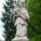 Zdjęcie nr 1: Figura św. Jana Nepomucena na rozbudowanej podstawie. Na trzech stopniach ustawiono sześcian zwężający się ku górze, składający się z postumentu z wolutami w dolnej części ustawionego na cokole i zwieńczonego gzymsem. Na ścianie frontowej na rozpostartej, zwieńczonej mitrą książęcą tkaninie złocona inskrypcja „PROSZĘ / O JEDNO PO/ZDROWIENIE / ZA DUSZE / ZMARŁYCH / A(NNO) D(OMINI) 1759” a poniżej data „R(OKU) 1901” ponad czaszką i piszczelą. Na podstawie, na cokole ustawiono figurę św. Jana Nepomucena w kontrapoście, z prawą nogą ugiętą w kolanie. Obiema rękami przytrzymuje krucyfiks i złotą palmę męczeństwa. Ma podłużną, smutną twarz o wklęsłych policzkach, niewielkich oczach, wąskim nosie i zamkniętych ustach okoloną krótką brodą i krótkimi włosami odwijającymi się do tyłu. Jest ubrany w strój kanonika: ma narzuconą na komżę i sutannę futrzaną almucję, a na głowie biret. Wokół głowy nimb z pięcioma złoconymi gwiazdkami. Na stopniu cokoło napis „ODNOWIŁA RADA GMINNA”. 