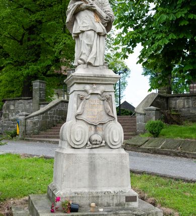 Zdjęcie nr 2: Figura św. Jana Nepomucena na rozbudowanej podstawie. Na trzech stopniach ustawiono sześcian zwężający się ku górze, składający się z postumentu z wolutami w dolnej części ustawionego na cokole i zwieńczonego gzymsem. Na ścianie frontowej na rozpostartej, zwieńczonej mitrą książęcą tkaninie złocona inskrypcja „PROSZĘ / O JEDNO PO/ZDROWIENIE / ZA DUSZE / ZMARŁYCH / A(NNO) D(OMINI) 1759” a poniżej data „R(OKU) 1901” ponad czaszką i piszczelą. Na podstawie, na cokole ustawiono figurę św. Jana Nepomucena w kontrapoście, z prawą nogą ugiętą w kolanie. Obiema rękami przytrzymuje krucyfiks i złotą palmę męczeństwa. Ma podłużną, smutną twarz o wklęsłych policzkach, niewielkich oczach, wąskim nosie i zamkniętych ustach okoloną krótką brodą i krótkimi włosami odwijającymi się do tyłu. Jest ubrany w strój kanonika: ma narzuconą na komżę i sutannę futrzaną almucję, a na głowie biret. Wokół głowy nimb z pięcioma złoconymi gwiazdkami. Na stopniu cokoło napis „ODNOWIŁA RADA GMINNA”. 