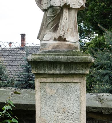 Zdjęcie nr 3: Na dwóch stopniach ustawiono wysoki cokół zwieńczony gzymsem, na którym na podstawie znajduje się figura św. Jana Nepomucena. Z czterech stron cokołu są fakturowane płyciny, na przedniej wyryto datę 1779. Święty o esowato wygiętej sylwetce, w kontrapoście, z prawą nogą zgiętą w kolanie. W obu rękach trzyma krucyfiks. Ma podłużną twarz okoloną krótkim zarostem i długimi, spływającymi na kark włosami. Ma półprzymknięte oczy, mały, wąski nos i rozciągnięte w delikatnym uśmiechu usta. Jest ubrany w strój kanonika: ma narzuconą na sutannę i komżę futrzaną almucję, a na głowie biret. Wokół głowy nimb z sześcioma gwiazdkami. 