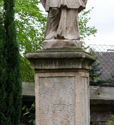 Zdjęcie nr 2: Na dwóch stopniach ustawiono wysoki cokół zwieńczony gzymsem, na którym na podstawie znajduje się figura św. Jana Nepomucena. Z czterech stron cokołu są fakturowane płyciny, na przedniej wyryto datę 1779. Święty o esowato wygiętej sylwetce, w kontrapoście, z prawą nogą zgiętą w kolanie. W obu rękach trzyma krucyfiks. Ma podłużną twarz okoloną krótkim zarostem i długimi, spływającymi na kark włosami. Ma półprzymknięte oczy, mały, wąski nos i rozciągnięte w delikatnym uśmiechu usta. Jest ubrany w strój kanonika: ma narzuconą na sutannę i komżę futrzaną almucję, a na głowie biret. Wokół głowy nimb z sześcioma gwiazdkami. 
