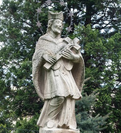 Zdjęcie nr 1: Na dwóch stopniach ustawiono wysoki cokół zwieńczony gzymsem, na którym na podstawie znajduje się figura św. Jana Nepomucena. Z czterech stron cokołu są fakturowane płyciny, na przedniej wyryto datę 1779. Święty o esowato wygiętej sylwetce, w kontrapoście, z prawą nogą zgiętą w kolanie. W obu rękach trzyma krucyfiks. Ma podłużną twarz okoloną krótkim zarostem i długimi, spływającymi na kark włosami. Ma półprzymknięte oczy, mały, wąski nos i rozciągnięte w delikatnym uśmiechu usta. Jest ubrany w strój kanonika: ma narzuconą na sutannę i komżę futrzaną almucję, a na głowie biret. Wokół głowy nimb z sześcioma gwiazdkami. 