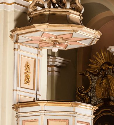 Zdjęcie nr 2: Ambona przyścienna składająca się z kosza, zaplecka, baldachimu i schodów z balustradą, zawieszona przy wschodnim filarze nawy. Kosz na planie sześcioboku, w narożach  ustawiono pilastry wsparte na cokołach, pomiędzy którymi prostokątne pola. Wszystkie płaszczyzny zdobione prostokątnymi płycinami dekorowanymi płaskorzeźbionymi postaciami ewangelistów oraz Chrystusa ukazanymi w półpostaci. Każdy z ewangelistów jest ubrany w długą złotą suknię i złoty płaszcz, trzyma w rękach otwartą księgę (św. Jan zwój), towarzyszy mu atrybut umożliwiający identyfikację. Chrystus jest ustawiony frontalnie, prawą ręką błogosławi, w lewej trzyma jabłko królewskie. Ma opadające na ramiona, zwinięte w pukle włosy oraz brodę. Podstawa kosza stożkowata, ujęta w narożach listwami, z pozłacaną szyszką od dołu. Zaplecek w kształcie prostokąta z płyciną ujętą pilastrami, wewnątrz której aplikowany stylizowany ornament roślinny. Baldachim powtarzający plan kosza, ujęty profilowanym gzymsem, wyłamanym w narożach, zwieńczony pełnoplastyczną figurą Chrystusa Zmartwychwstałego trzymającego krzyż, ustawioną na wielobocznej podstawie wspartej na sześciu wolutach. Chrystus został ukazany w całej postaci, z prawą ręką uniesioną w geście błogosławieństwa, lewą przytrzymującą krzyż. Ma owalną twarz okoloną długimi włosami, które spływają mu na ramiona i plecy; nad głową okrągły nimb. Jest ubrany w długą suknię, na którą ma zarzucony płaszcz spięty na ramionach. Obie szaty łamią się ostrymi fałdami. W podniebiu baldachimu znajduje się podwójna, ośmioramienna gwiazda wydzielona pozłacanymi listwami, z lampą na środku. Balustrada z profilowaną poręczą, artykułowana romboidalnymi płycinami i pilastrami. Kolorystyka pastelowa, jasna. Struktura w kolorze jasnoszarym, płyciny jasnobeżowe, detal architektoniczny, ornamenty oraz szaty pozłacane, atrybuty ewangelistów posrebrzane.