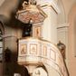 Zdjęcie nr 1: Ambona przyścienna składająca się z kosza, zaplecka, baldachimu i schodów z balustradą, zawieszona przy wschodnim filarze nawy. Kosz na planie sześcioboku, w narożach  ustawiono pilastry wsparte na cokołach, pomiędzy którymi prostokątne pola. Wszystkie płaszczyzny zdobione prostokątnymi płycinami dekorowanymi płaskorzeźbionymi postaciami ewangelistów oraz Chrystusa ukazanymi w półpostaci. Każdy z ewangelistów jest ubrany w długą złotą suknię i złoty płaszcz, trzyma w rękach otwartą księgę (św. Jan zwój), towarzyszy mu atrybut umożliwiający identyfikację. Chrystus jest ustawiony frontalnie, prawą ręką błogosławi, w lewej trzyma jabłko królewskie. Ma opadające na ramiona, zwinięte w pukle włosy oraz brodę. Podstawa kosza stożkowata, ujęta w narożach listwami, z pozłacaną szyszką od dołu. Zaplecek w kształcie prostokąta z płyciną ujętą pilastrami, wewnątrz której aplikowany stylizowany ornament roślinny. Baldachim powtarzający plan kosza, ujęty profilowanym gzymsem, wyłamanym w narożach, zwieńczony pełnoplastyczną figurą Chrystusa Zmartwychwstałego trzymającego krzyż, ustawioną na wielobocznej podstawie wspartej na sześciu wolutach. Chrystus został ukazany w całej postaci, z prawą ręką uniesioną w geście błogosławieństwa, lewą przytrzymującą krzyż. Ma owalną twarz okoloną długimi włosami, które spływają mu na ramiona i plecy; nad głową okrągły nimb. Jest ubrany w długą suknię, na którą ma zarzucony płaszcz spięty na ramionach. Obie szaty łamią się ostrymi fałdami. W podniebiu baldachimu znajduje się podwójna, ośmioramienna gwiazda wydzielona pozłacanymi listwami, z lampą na środku. Balustrada z profilowaną poręczą, artykułowana romboidalnymi płycinami i pilastrami. Kolorystyka pastelowa, jasna. Struktura w kolorze jasnoszarym, płyciny jasnobeżowe, detal architektoniczny, ornamenty oraz szaty pozłacane, atrybuty ewangelistów posrebrzane.