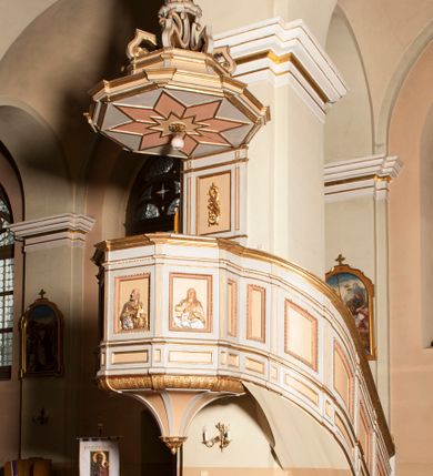 Zdjęcie nr 1: Ambona przyścienna składająca się z kosza, zaplecka, baldachimu i schodów z balustradą, zawieszona przy wschodnim filarze nawy. Kosz na planie sześcioboku, w narożach  ustawiono pilastry wsparte na cokołach, pomiędzy którymi prostokątne pola. Wszystkie płaszczyzny zdobione prostokątnymi płycinami dekorowanymi płaskorzeźbionymi postaciami ewangelistów oraz Chrystusa ukazanymi w półpostaci. Każdy z ewangelistów jest ubrany w długą złotą suknię i złoty płaszcz, trzyma w rękach otwartą księgę (św. Jan zwój), towarzyszy mu atrybut umożliwiający identyfikację. Chrystus jest ustawiony frontalnie, prawą ręką błogosławi, w lewej trzyma jabłko królewskie. Ma opadające na ramiona, zwinięte w pukle włosy oraz brodę. Podstawa kosza stożkowata, ujęta w narożach listwami, z pozłacaną szyszką od dołu. Zaplecek w kształcie prostokąta z płyciną ujętą pilastrami, wewnątrz której aplikowany stylizowany ornament roślinny. Baldachim powtarzający plan kosza, ujęty profilowanym gzymsem, wyłamanym w narożach, zwieńczony pełnoplastyczną figurą Chrystusa Zmartwychwstałego trzymającego krzyż, ustawioną na wielobocznej podstawie wspartej na sześciu wolutach. Chrystus został ukazany w całej postaci, z prawą ręką uniesioną w geście błogosławieństwa, lewą przytrzymującą krzyż. Ma owalną twarz okoloną długimi włosami, które spływają mu na ramiona i plecy; nad głową okrągły nimb. Jest ubrany w długą suknię, na którą ma zarzucony płaszcz spięty na ramionach. Obie szaty łamią się ostrymi fałdami. W podniebiu baldachimu znajduje się podwójna, ośmioramienna gwiazda wydzielona pozłacanymi listwami, z lampą na środku. Balustrada z profilowaną poręczą, artykułowana romboidalnymi płycinami i pilastrami. Kolorystyka pastelowa, jasna. Struktura w kolorze jasnoszarym, płyciny jasnobeżowe, detal architektoniczny, ornamenty oraz szaty pozłacane, atrybuty ewangelistów posrebrzane.