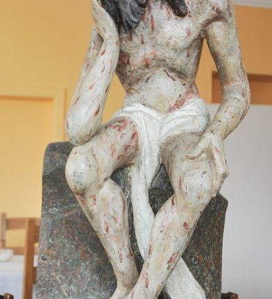 Zdjęcie nr 2: Rzeźba pełna, całopostaciowa, ustawiona na niewysokim cokole. Chrystus ukazany w pozycji siedzącej, na podstawie imitującej blok skalny. Prawą ręką, opartą na udzie  podtrzymuje głowę, lewą dłoń opiera na lewej nodze; kolana ma rozchylone, lewą stopę ustawioną przed prawą. Chrystus ma pociągłą twarz o wydatnych kościach policzkowych, wąskie brwi opadające prostą linią w dół, długi nos, zagłębione oczy i wyraźnie zaznaczone usta. Długie włosy opadają mu na ramiona i plecy, broda dzieli się w dwa pukle; na głowie grube zwoje korony cierniowej.  Biodra ma przepasane białym perizonium, którego długi, wolny koniec opada między udami i za lewą nogą aż na podłoże. Na całym ciele zaznaczono ślady męki – równomiernie rozmieszczone krople spływającej krwi. W partiach ciała polichromia naturalistyczna.