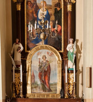 Zdjęcie nr 3: Obraz w kształcie stojącego prostokąta zamkniętego półkoliście. Pośrodku, na tle pejzażu z rozległym szaro-błękitnym niebem przedstawiona św. Barbara, ukazana w całej postaci, frontalnie, w pozycji stojącej, z głową lekko uniesioną. Prawą, zgiętą i uniesioną ręką trzyma kielich z hostią, lewą, opuszczoną przytrzymuje miecz i palmę męczeństwa. Święta ma twarz o dziewczęcych rysach i naturalnej karnacji okoloną ciemnymi, upiętymi włosami. Wzrok kieruje ku górze, nos ma prosty, usta niewielkie. Na głowie korona otwarta, wokół świetlisty nimb. Święta ubrana jest w bogato zdobioną, długą zieloną suknię z gorsetem, owalnym dekoltem i bufiastymi rękawami oraz czerwony płaszcz z ornamentalną bordiurą okrywający ramiona i nogi. Nad głową świętej widoczne uskrzydlone główki anielskie, w oddali po lewej stronie murowana z cegły wieża z machikułami nakryta dachem stożkowym.