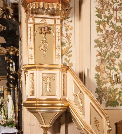 Zdjęcie nr 5: Ambona przyścienna składająca się z kosza, zaplecka, baldachimu i schodów z balustradą, zawieszona na lewej ścianie prezbiterium. Kosz na planie ośmioboku, o ściankach naprzemiennie prostych i wklęsłych, dekorowanych płycinami z plakietami ornamentalnymi. Podstawa kosza stożkowa, z wypełniającymi płaszczyzny płycinami, z pozłacaną szyszką u dołu. Zaplecek w kształcie prostokąta z prostokątną płyciną, wewnątrz której aplikowany stylizowany ornament roślinny i dwie główki anielskie poniżej łuku trójlistnego. Baldachim powtarzający plan kosza, ujęty profilowanym gzymsem, wyłamanym w narożach, z podwieszonym lambrekinem i zwieńczony rzędem ornamentu złożonego ze stylizowanych lilii.  Baldachim wieńczy pełnoplastyczna figura Chrystusa Zmartwychwstałego ustawiona na stożkowej podstawie. Chrystus został ukazany w całej postaci, z prawą ręką uniesioną w geście błogosławieństwa, lewą przytrzymując płaszcz. Ma owalną twarz okoloną długimi włosami, które spływają mu na ramiona i plecy. Wokół bioder ma obwiązane białe perizonium, na plecach zarzucony złoty płaszcz spięty na dekolcie; szaty łamią się ostrymi fałdami. W podniebiu baldachimu znajduje się rzeźba srebrnej gołębicy Ducha Świętego. Balustrada z profilowaną poręczą, artykułowana romboidalnymi płycinami  z plakietami. Kolorystyka pastelowa, jasna. Struktura w kolorze kremowym, płyciny jasnoszare, fryz baldachimu bordowy detal architektoniczny i ornamenty pozłacane. 