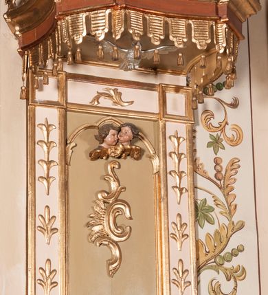 Zdjęcie nr 1: Ambona przyścienna składająca się z kosza, zaplecka, baldachimu i schodów z balustradą, zawieszona na lewej ścianie prezbiterium. Kosz na planie ośmioboku, o ściankach naprzemiennie prostych i wklęsłych, dekorowanych płycinami z plakietami ornamentalnymi. Podstawa kosza stożkowa, z wypełniającymi płaszczyzny płycinami, z pozłacaną szyszką u dołu. Zaplecek w kształcie prostokąta z prostokątną płyciną, wewnątrz której aplikowany stylizowany ornament roślinny i dwie główki anielskie poniżej łuku trójlistnego. Baldachim powtarzający plan kosza, ujęty profilowanym gzymsem, wyłamanym w narożach, z podwieszonym lambrekinem i zwieńczony rzędem ornamentu złożonego ze stylizowanych lilii.  Baldachim wieńczy pełnoplastyczna figura Chrystusa Zmartwychwstałego ustawiona na stożkowej podstawie. Chrystus został ukazany w całej postaci, z prawą ręką uniesioną w geście błogosławieństwa, lewą przytrzymując płaszcz. Ma owalną twarz okoloną długimi włosami, które spływają mu na ramiona i plecy. Wokół bioder ma obwiązane białe perizonium, na plecach zarzucony złoty płaszcz spięty na dekolcie; szaty łamią się ostrymi fałdami. W podniebiu baldachimu znajduje się rzeźba srebrnej gołębicy Ducha Świętego. Balustrada z profilowaną poręczą, artykułowana romboidalnymi płycinami  z plakietami. Kolorystyka pastelowa, jasna. Struktura w kolorze kremowym, płyciny jasnoszare, fryz baldachimu bordowy detal architektoniczny i ornamenty pozłacane. 