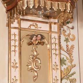 Zdjęcie nr 1: Ambona przyścienna składająca się z kosza, zaplecka, baldachimu i schodów z balustradą, zawieszona na lewej ścianie prezbiterium. Kosz na planie ośmioboku, o ściankach naprzemiennie prostych i wklęsłych, dekorowanych płycinami z plakietami ornamentalnymi. Podstawa kosza stożkowa, z wypełniającymi płaszczyzny płycinami, z pozłacaną szyszką u dołu. Zaplecek w kształcie prostokąta z prostokątną płyciną, wewnątrz której aplikowany stylizowany ornament roślinny i dwie główki anielskie poniżej łuku trójlistnego. Baldachim powtarzający plan kosza, ujęty profilowanym gzymsem, wyłamanym w narożach, z podwieszonym lambrekinem i zwieńczony rzędem ornamentu złożonego ze stylizowanych lilii.  Baldachim wieńczy pełnoplastyczna figura Chrystusa Zmartwychwstałego ustawiona na stożkowej podstawie. Chrystus został ukazany w całej postaci, z prawą ręką uniesioną w geście błogosławieństwa, lewą przytrzymując płaszcz. Ma owalną twarz okoloną długimi włosami, które spływają mu na ramiona i plecy. Wokół bioder ma obwiązane białe perizonium, na plecach zarzucony złoty płaszcz spięty na dekolcie; szaty łamią się ostrymi fałdami. W podniebiu baldachimu znajduje się rzeźba srebrnej gołębicy Ducha Świętego. Balustrada z profilowaną poręczą, artykułowana romboidalnymi płycinami  z plakietami. Kolorystyka pastelowa, jasna. Struktura w kolorze kremowym, płyciny jasnoszare, fryz baldachimu bordowy detal architektoniczny i ornamenty pozłacane. 