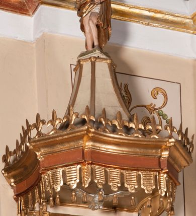 Zdjęcie nr 3: Ambona przyścienna składająca się z kosza, zaplecka, baldachimu i schodów z balustradą, zawieszona na lewej ścianie prezbiterium. Kosz na planie ośmioboku, o ściankach naprzemiennie prostych i wklęsłych, dekorowanych płycinami z plakietami ornamentalnymi. Podstawa kosza stożkowa, z wypełniającymi płaszczyzny płycinami, z pozłacaną szyszką u dołu. Zaplecek w kształcie prostokąta z prostokątną płyciną, wewnątrz której aplikowany stylizowany ornament roślinny i dwie główki anielskie poniżej łuku trójlistnego. Baldachim powtarzający plan kosza, ujęty profilowanym gzymsem, wyłamanym w narożach, z podwieszonym lambrekinem i zwieńczony rzędem ornamentu złożonego ze stylizowanych lilii.  Baldachim wieńczy pełnoplastyczna figura Chrystusa Zmartwychwstałego ustawiona na stożkowej podstawie. Chrystus został ukazany w całej postaci, z prawą ręką uniesioną w geście błogosławieństwa, lewą przytrzymując płaszcz. Ma owalną twarz okoloną długimi włosami, które spływają mu na ramiona i plecy. Wokół bioder ma obwiązane białe perizonium, na plecach zarzucony złoty płaszcz spięty na dekolcie; szaty łamią się ostrymi fałdami. W podniebiu baldachimu znajduje się rzeźba srebrnej gołębicy Ducha Świętego. Balustrada z profilowaną poręczą, artykułowana romboidalnymi płycinami  z plakietami. Kolorystyka pastelowa, jasna. Struktura w kolorze kremowym, płyciny jasnoszare, fryz baldachimu bordowy detal architektoniczny i ornamenty pozłacane. 