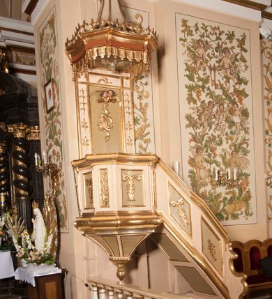 Zdjęcie nr 4: Ambona przyścienna składająca się z kosza, zaplecka, baldachimu i schodów z balustradą, zawieszona na lewej ścianie prezbiterium. Kosz na planie ośmioboku, o ściankach naprzemiennie prostych i wklęsłych, dekorowanych płycinami z plakietami ornamentalnymi. Podstawa kosza stożkowa, z wypełniającymi płaszczyzny płycinami, z pozłacaną szyszką u dołu. Zaplecek w kształcie prostokąta z prostokątną płyciną, wewnątrz której aplikowany stylizowany ornament roślinny i dwie główki anielskie poniżej łuku trójlistnego. Baldachim powtarzający plan kosza, ujęty profilowanym gzymsem, wyłamanym w narożach, z podwieszonym lambrekinem i zwieńczony rzędem ornamentu złożonego ze stylizowanych lilii.  Baldachim wieńczy pełnoplastyczna figura Chrystusa Zmartwychwstałego ustawiona na stożkowej podstawie. Chrystus został ukazany w całej postaci, z prawą ręką uniesioną w geście błogosławieństwa, lewą przytrzymując płaszcz. Ma owalną twarz okoloną długimi włosami, które spływają mu na ramiona i plecy. Wokół bioder ma obwiązane białe perizonium, na plecach zarzucony złoty płaszcz spięty na dekolcie; szaty łamią się ostrymi fałdami. W podniebiu baldachimu znajduje się rzeźba srebrnej gołębicy Ducha Świętego. Balustrada z profilowaną poręczą, artykułowana romboidalnymi płycinami  z plakietami. Kolorystyka pastelowa, jasna. Struktura w kolorze kremowym, płyciny jasnoszare, fryz baldachimu bordowy detal architektoniczny i ornamenty pozłacane. 
