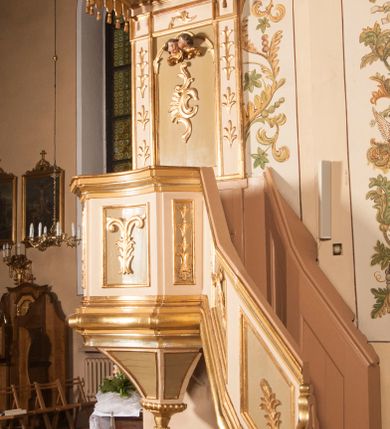 Zdjęcie nr 6: Ambona przyścienna składająca się z kosza, zaplecka, baldachimu i schodów z balustradą, zawieszona na lewej ścianie prezbiterium. Kosz na planie ośmioboku, o ściankach naprzemiennie prostych i wklęsłych, dekorowanych płycinami z plakietami ornamentalnymi. Podstawa kosza stożkowa, z wypełniającymi płaszczyzny płycinami, z pozłacaną szyszką u dołu. Zaplecek w kształcie prostokąta z prostokątną płyciną, wewnątrz której aplikowany stylizowany ornament roślinny i dwie główki anielskie poniżej łuku trójlistnego. Baldachim powtarzający plan kosza, ujęty profilowanym gzymsem, wyłamanym w narożach, z podwieszonym lambrekinem i zwieńczony rzędem ornamentu złożonego ze stylizowanych lilii.  Baldachim wieńczy pełnoplastyczna figura Chrystusa Zmartwychwstałego ustawiona na stożkowej podstawie. Chrystus został ukazany w całej postaci, z prawą ręką uniesioną w geście błogosławieństwa, lewą przytrzymując płaszcz. Ma owalną twarz okoloną długimi włosami, które spływają mu na ramiona i plecy. Wokół bioder ma obwiązane białe perizonium, na plecach zarzucony złoty płaszcz spięty na dekolcie; szaty łamią się ostrymi fałdami. W podniebiu baldachimu znajduje się rzeźba srebrnej gołębicy Ducha Świętego. Balustrada z profilowaną poręczą, artykułowana romboidalnymi płycinami  z plakietami. Kolorystyka pastelowa, jasna. Struktura w kolorze kremowym, płyciny jasnoszare, fryz baldachimu bordowy detal architektoniczny i ornamenty pozłacane. 