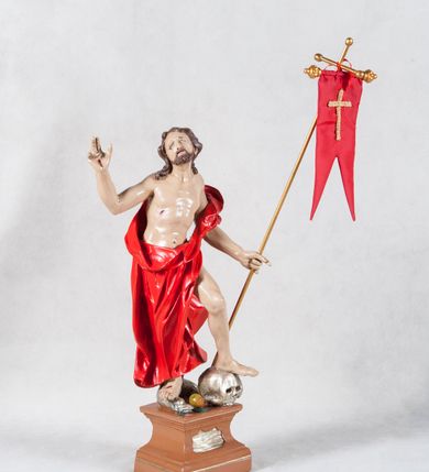 Zdjęcie nr 2: Rzeźba Chrystusa Zmartwychwstałego pełna, ustawiona na wysokim, profilowanym cokoliku. Chrystus w ujęciu całopostaciowym, przedstawiony z krzyżem i proporcem, zwrócony lekko w prawo. Ujęty w pozycji stojącej w kontrapoście z ugiętą lewą nogą wspartą na czaszce Adama. Prawą depcze węża trzymającego w pysku jabłko. Twarz owalna, okolona zarostem, włosy długie, zbite w pukle, luźno opadają na ramiona. Na plecy narzucony płaszcz, silnie drapowany na linii bioder i osłaniający prawą nogę. Stopy bose. Na boku korpusu, dłoniach oraz stopach widoczne rany. Jezus prawą rękę ma uniesioną w geście błogosławieństwa, lewą podtrzymuje krzyż i proporzec. 