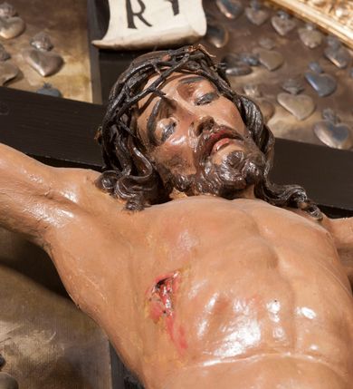Zdjęcie nr 5: Wizerunek Chrystusa przybitego trzema gwoździami do krzyża łacińskiego na tle obrazu z przedstawieniem widoku Jerozolimy. Ciało wyprostowane, ręce rozciągnięte do boku, barki uniesione, nogi delikatnie ugięte w kolanach, prawe powyżej lewego. Głowa opada na prawy bark. Jezus ma podłużną twarz, usta i oczy zamknięte, nos prosty; okala ją dzieląca się w pukle broda oraz zwijające się w fale włosy opadające na plecy i barki. Na głowie cienka, ciemnobrązowa korona cierniowa. Biodra otacza złote, drobno marszczone perizonium odwijające się i przytrzymane sznurem związanym na prawym boku.  W miejscach przebić gwoździami oraz w  ranie w boku ślady krwi. Krzyż gładki, na górnej belce titulus w formie zwoju z literami „IN/RI” powyżej wotum złożonego z dwóch serc. W tle obraz z płaskorzeźbionym przedstawieniem wzgórz i sylwetki miasta oraz malowanym, ciemnobrunatnym tłem rozjaśnionym po lewej stronie Chrystusa i na skrzyżowaniu belek krzyża. Polichromia w partiach ciała naturalistyczna, krzyż ciemnobrązowy. Do tła przymocowano liczne dary wotywne. Polichromia w partiach ciała naturalistyczna.