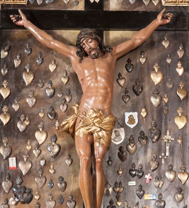 Zdjęcie nr 4: Wizerunek Chrystusa przybitego trzema gwoździami do krzyża łacińskiego na tle obrazu z przedstawieniem widoku Jerozolimy. Ciało wyprostowane, ręce rozciągnięte do boku, barki uniesione, nogi delikatnie ugięte w kolanach, prawe powyżej lewego. Głowa opada na prawy bark. Jezus ma podłużną twarz, usta i oczy zamknięte, nos prosty; okala ją dzieląca się w pukle broda oraz zwijające się w fale włosy opadające na plecy i barki. Na głowie cienka, ciemnobrązowa korona cierniowa. Biodra otacza złote, drobno marszczone perizonium odwijające się i przytrzymane sznurem związanym na prawym boku.  W miejscach przebić gwoździami oraz w  ranie w boku ślady krwi. Krzyż gładki, na górnej belce titulus w formie zwoju z literami „IN/RI” powyżej wotum złożonego z dwóch serc. W tle obraz z płaskorzeźbionym przedstawieniem wzgórz i sylwetki miasta oraz malowanym, ciemnobrunatnym tłem rozjaśnionym po lewej stronie Chrystusa i na skrzyżowaniu belek krzyża. Polichromia w partiach ciała naturalistyczna, krzyż ciemnobrązowy. Do tła przymocowano liczne dary wotywne. Polichromia w partiach ciała naturalistyczna.