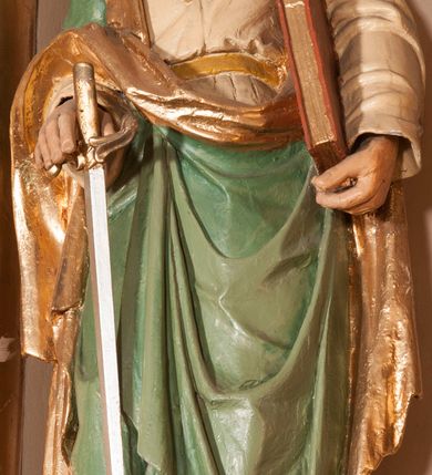 Zdjęcie nr 2: Rzeźba drążona na cokole przedstawiająca św. Pawła ukazanego w całej postaci, zwróconego nieznacznie w lewo. Święty prawą, zgiętą w łokciu ręką trzyma rękojeść opartego o ziemię miecza, lewą podtrzymuje zamkniętą księgę. Ma owalną twarz z dużymi, szeroko otwartymi oczami, prostym nosem oraz wydatnymi ustami; okalają ją ciemnobrązowe, zaczesane do tyłu włosy. Święty ma brodę sięgającą piersi oraz wąsy. Jest ubrany w kremową tunikę z długimi rękawami, zapinaną na guziki i przepasaną w talii oraz zielony płaszcz ze złotym podbiciem przerzucony przez prawe ramię, owijający się wokół bioder i przełożony przez prawą rękę. Tkanina płaszcza spływa do ziemi kaskadowymi fałdami. 
W partiach ciała polichromia naturalistyczna, płaszcz i atrybuty złocone i srebrzone.
