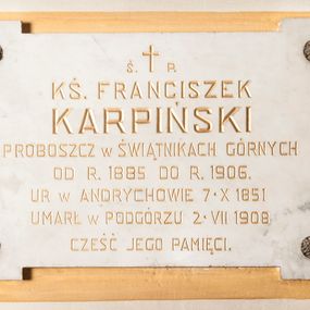 Zdjęcie nr 1: Wmurowana w ścianę kościoła tablica epitafijna w formie leżącego prostokąta z uszakami, na środku których umieszczono metalowe rozetki maskujące miejsca mocowania. Na środku tablicy widnieje złocona inskrypcja „Ś(WIĘTEJ) P(AMIĘCI) / KS(IĄDZ) FRANCISZEK / KARPIŃSKI / PROBOSZCZ W ŚWIĄTNIKACH GÓRNYCH / OD R(OKU) 1885 DO R(OKU) 1906. / UR[ODZONY] W ANDRYCHOWIE 7. X 1851 / UMARŁ W PODGÓRZU 2. VII 1908 / CZEŚĆ JEGO PAMIĘCI”. 