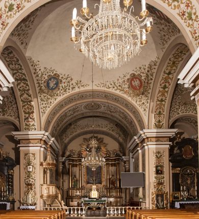 Zdjęcie nr 5: Kościół murowany, trójnawowy, halowy, na rzucie prostokąta z wydłużonym, skierowanym na północ prezbiterium z dobudowaną po zachodniej stronie zakrystią. Prezbiterium jednoprzęsłowe, węższe od korpusu, zamknięte prosto. Nawa główna oraz boczne dwuprzęsłowe, rozdzielone masywnymi opilastrowanymi filarami; artykulacja ścian przy pomocy filarów. Sklepienie prezbiterium kolebkowe, naw żagielkowe, przedzielone gurtami. Od południa chór muzyczny mieszczący się między pierwszymi filarami i powyżej kruchty wejściowej. W prezbiterium jedno okno od wschodu, drugie w zakrystii, w nawach bocznych po dwa. Główne wejście od południa flankowane przez dwa boczne, kolejne do zakrystii i między zakrystią i prezbiterium. Niektóre okna wypełnione gomułkami, ściany i sklepienia kościoła pokrywa polichromia utworzona z dekoracji floralnych i umieszczonych w jej obrębie symboli ewangelistów, herbów i putt wykonujących prace kojarzące się z regionem (bartnicze, ślusarskie, kowalskie itd.). Fasada dwuwieżowa, trójosiowa, dwuprzęsłowa, z portalem na każdej osi. Podziały pionowe przeprowadzono za pomocą półkolumienek i lizen, poziome w formie fryzu kostkowego wyniesionego nad środkowym portalem. Drzwi osi głównej w kształcie stojącego prostokąta ujętego półkolumnami dźwigającymi archiwoltę utworzoną z trzech łuków i trójkątny przyczółek. Drzwi osi bocznych zwieńczone przyczółkami. W drugiej kondygnacji na osi okulus i półokrągła konchowa wnęka z figurą Najświętszego Serca Jezusa poniżej trójkątnego szczytu dekorowanego fryzem arkadkowym. W osiach bocznych okna w kształcie stojącego prostokąta zamkniętego półkoliście. Wyżej dwie kondygnacje wież oddzielone gzymsem, w pierwszej  umieszczono okna, w drugiej tarcze zegarów. Elewacje boczne gładkie, przeprute jedynie oknami. Nawy przykrywa dach dwuspadowy, wieże namiotowe z fartuchem zwieńczone krzyżami.