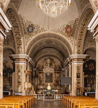 Zdjęcie nr 6: Kościół murowany, trójnawowy, halowy, na rzucie prostokąta z wydłużonym, skierowanym na północ prezbiterium z dobudowaną po zachodniej stronie zakrystią. Prezbiterium jednoprzęsłowe, węższe od korpusu, zamknięte prosto. Nawa główna oraz boczne dwuprzęsłowe, rozdzielone masywnymi opilastrowanymi filarami; artykulacja ścian przy pomocy filarów. Sklepienie prezbiterium kolebkowe, naw żagielkowe, przedzielone gurtami. Od południa chór muzyczny mieszczący się między pierwszymi filarami i powyżej kruchty wejściowej. W prezbiterium jedno okno od wschodu, drugie w zakrystii, w nawach bocznych po dwa. Główne wejście od południa flankowane przez dwa boczne, kolejne do zakrystii i między zakrystią i prezbiterium. Niektóre okna wypełnione gomułkami, ściany i sklepienia kościoła pokrywa polichromia utworzona z dekoracji floralnych i umieszczonych w jej obrębie symboli ewangelistów, herbów i putt wykonujących prace kojarzące się z regionem (bartnicze, ślusarskie, kowalskie itd.). Fasada dwuwieżowa, trójosiowa, dwuprzęsłowa, z portalem na każdej osi. Podziały pionowe przeprowadzono za pomocą półkolumienek i lizen, poziome w formie fryzu kostkowego wyniesionego nad środkowym portalem. Drzwi osi głównej w kształcie stojącego prostokąta ujętego półkolumnami dźwigającymi archiwoltę utworzoną z trzech łuków i trójkątny przyczółek. Drzwi osi bocznych zwieńczone przyczółkami. W drugiej kondygnacji na osi okulus i półokrągła konchowa wnęka z figurą Najświętszego Serca Jezusa poniżej trójkątnego szczytu dekorowanego fryzem arkadkowym. W osiach bocznych okna w kształcie stojącego prostokąta zamkniętego półkoliście. Wyżej dwie kondygnacje wież oddzielone gzymsem, w pierwszej  umieszczono okna, w drugiej tarcze zegarów. Elewacje boczne gładkie, przeprute jedynie oknami. Nawy przykrywa dach dwuspadowy, wieże namiotowe z fartuchem zwieńczone krzyżami.