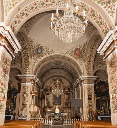 Zdjęcie nr 9: Kościół murowany, trójnawowy, halowy, na rzucie prostokąta z wydłużonym, skierowanym na północ prezbiterium z dobudowaną po zachodniej stronie zakrystią. Prezbiterium jednoprzęsłowe, węższe od korpusu, zamknięte prosto. Nawa główna oraz boczne dwuprzęsłowe, rozdzielone masywnymi opilastrowanymi filarami; artykulacja ścian przy pomocy filarów. Sklepienie prezbiterium kolebkowe, naw żagielkowe, przedzielone gurtami. Od południa chór muzyczny mieszczący się między pierwszymi filarami i powyżej kruchty wejściowej. W prezbiterium jedno okno od wschodu, drugie w zakrystii, w nawach bocznych po dwa. Główne wejście od południa flankowane przez dwa boczne, kolejne do zakrystii i między zakrystią i prezbiterium. Niektóre okna wypełnione gomułkami, ściany i sklepienia kościoła pokrywa polichromia utworzona z dekoracji floralnych i umieszczonych w jej obrębie symboli ewangelistów, herbów i putt wykonujących prace kojarzące się z regionem (bartnicze, ślusarskie, kowalskie itd.). Fasada dwuwieżowa, trójosiowa, dwuprzęsłowa, z portalem na każdej osi. Podziały pionowe przeprowadzono za pomocą półkolumienek i lizen, poziome w formie fryzu kostkowego wyniesionego nad środkowym portalem. Drzwi osi głównej w kształcie stojącego prostokąta ujętego półkolumnami dźwigającymi archiwoltę utworzoną z trzech łuków i trójkątny przyczółek. Drzwi osi bocznych zwieńczone przyczółkami. W drugiej kondygnacji na osi okulus i półokrągła konchowa wnęka z figurą Najświętszego Serca Jezusa poniżej trójkątnego szczytu dekorowanego fryzem arkadkowym. W osiach bocznych okna w kształcie stojącego prostokąta zamkniętego półkoliście. Wyżej dwie kondygnacje wież oddzielone gzymsem, w pierwszej  umieszczono okna, w drugiej tarcze zegarów. Elewacje boczne gładkie, przeprute jedynie oknami. Nawy przykrywa dach dwuspadowy, wieże namiotowe z fartuchem zwieńczone krzyżami.