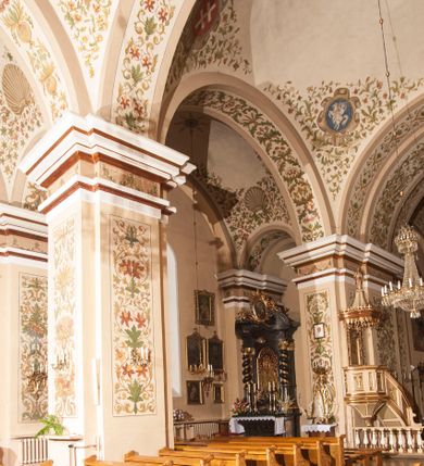 Zdjęcie nr 10: Kościół murowany, trójnawowy, halowy, na rzucie prostokąta z wydłużonym, skierowanym na północ prezbiterium z dobudowaną po zachodniej stronie zakrystią. Prezbiterium jednoprzęsłowe, węższe od korpusu, zamknięte prosto. Nawa główna oraz boczne dwuprzęsłowe, rozdzielone masywnymi opilastrowanymi filarami; artykulacja ścian przy pomocy filarów. Sklepienie prezbiterium kolebkowe, naw żagielkowe, przedzielone gurtami. Od południa chór muzyczny mieszczący się między pierwszymi filarami i powyżej kruchty wejściowej. W prezbiterium jedno okno od wschodu, drugie w zakrystii, w nawach bocznych po dwa. Główne wejście od południa flankowane przez dwa boczne, kolejne do zakrystii i między zakrystią i prezbiterium. Niektóre okna wypełnione gomułkami, ściany i sklepienia kościoła pokrywa polichromia utworzona z dekoracji floralnych i umieszczonych w jej obrębie symboli ewangelistów, herbów i putt wykonujących prace kojarzące się z regionem (bartnicze, ślusarskie, kowalskie itd.). Fasada dwuwieżowa, trójosiowa, dwuprzęsłowa, z portalem na każdej osi. Podziały pionowe przeprowadzono za pomocą półkolumienek i lizen, poziome w formie fryzu kostkowego wyniesionego nad środkowym portalem. Drzwi osi głównej w kształcie stojącego prostokąta ujętego półkolumnami dźwigającymi archiwoltę utworzoną z trzech łuków i trójkątny przyczółek. Drzwi osi bocznych zwieńczone przyczółkami. W drugiej kondygnacji na osi okulus i półokrągła konchowa wnęka z figurą Najświętszego Serca Jezusa poniżej trójkątnego szczytu dekorowanego fryzem arkadkowym. W osiach bocznych okna w kształcie stojącego prostokąta zamkniętego półkoliście. Wyżej dwie kondygnacje wież oddzielone gzymsem, w pierwszej  umieszczono okna, w drugiej tarcze zegarów. Elewacje boczne gładkie, przeprute jedynie oknami. Nawy przykrywa dach dwuspadowy, wieże namiotowe z fartuchem zwieńczone krzyżami.