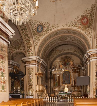 Zdjęcie nr 8: Kościół murowany, trójnawowy, halowy, na rzucie prostokąta z wydłużonym, skierowanym na północ prezbiterium z dobudowaną po zachodniej stronie zakrystią. Prezbiterium jednoprzęsłowe, węższe od korpusu, zamknięte prosto. Nawa główna oraz boczne dwuprzęsłowe, rozdzielone masywnymi opilastrowanymi filarami; artykulacja ścian przy pomocy filarów. Sklepienie prezbiterium kolebkowe, naw żagielkowe, przedzielone gurtami. Od południa chór muzyczny mieszczący się między pierwszymi filarami i powyżej kruchty wejściowej. W prezbiterium jedno okno od wschodu, drugie w zakrystii, w nawach bocznych po dwa. Główne wejście od południa flankowane przez dwa boczne, kolejne do zakrystii i między zakrystią i prezbiterium. Niektóre okna wypełnione gomułkami, ściany i sklepienia kościoła pokrywa polichromia utworzona z dekoracji floralnych i umieszczonych w jej obrębie symboli ewangelistów, herbów i putt wykonujących prace kojarzące się z regionem (bartnicze, ślusarskie, kowalskie itd.). Fasada dwuwieżowa, trójosiowa, dwuprzęsłowa, z portalem na każdej osi. Podziały pionowe przeprowadzono za pomocą półkolumienek i lizen, poziome w formie fryzu kostkowego wyniesionego nad środkowym portalem. Drzwi osi głównej w kształcie stojącego prostokąta ujętego półkolumnami dźwigającymi archiwoltę utworzoną z trzech łuków i trójkątny przyczółek. Drzwi osi bocznych zwieńczone przyczółkami. W drugiej kondygnacji na osi okulus i półokrągła konchowa wnęka z figurą Najświętszego Serca Jezusa poniżej trójkątnego szczytu dekorowanego fryzem arkadkowym. W osiach bocznych okna w kształcie stojącego prostokąta zamkniętego półkoliście. Wyżej dwie kondygnacje wież oddzielone gzymsem, w pierwszej  umieszczono okna, w drugiej tarcze zegarów. Elewacje boczne gładkie, przeprute jedynie oknami. Nawy przykrywa dach dwuspadowy, wieże namiotowe z fartuchem zwieńczone krzyżami.