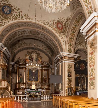 Zdjęcie nr 4: Kościół murowany, trójnawowy, halowy, na rzucie prostokąta z wydłużonym, skierowanym na północ prezbiterium z dobudowaną po zachodniej stronie zakrystią. Prezbiterium jednoprzęsłowe, węższe od korpusu, zamknięte prosto. Nawa główna oraz boczne dwuprzęsłowe, rozdzielone masywnymi opilastrowanymi filarami; artykulacja ścian przy pomocy filarów. Sklepienie prezbiterium kolebkowe, naw żagielkowe, przedzielone gurtami. Od południa chór muzyczny mieszczący się między pierwszymi filarami i powyżej kruchty wejściowej. W prezbiterium jedno okno od wschodu, drugie w zakrystii, w nawach bocznych po dwa. Główne wejście od południa flankowane przez dwa boczne, kolejne do zakrystii i między zakrystią i prezbiterium. Niektóre okna wypełnione gomułkami, ściany i sklepienia kościoła pokrywa polichromia utworzona z dekoracji floralnych i umieszczonych w jej obrębie symboli ewangelistów, herbów i putt wykonujących prace kojarzące się z regionem (bartnicze, ślusarskie, kowalskie itd.). Fasada dwuwieżowa, trójosiowa, dwuprzęsłowa, z portalem na każdej osi. Podziały pionowe przeprowadzono za pomocą półkolumienek i lizen, poziome w formie fryzu kostkowego wyniesionego nad środkowym portalem. Drzwi osi głównej w kształcie stojącego prostokąta ujętego półkolumnami dźwigającymi archiwoltę utworzoną z trzech łuków i trójkątny przyczółek. Drzwi osi bocznych zwieńczone przyczółkami. W drugiej kondygnacji na osi okulus i półokrągła konchowa wnęka z figurą Najświętszego Serca Jezusa poniżej trójkątnego szczytu dekorowanego fryzem arkadkowym. W osiach bocznych okna w kształcie stojącego prostokąta zamkniętego półkoliście. Wyżej dwie kondygnacje wież oddzielone gzymsem, w pierwszej  umieszczono okna, w drugiej tarcze zegarów. Elewacje boczne gładkie, przeprute jedynie oknami. Nawy przykrywa dach dwuspadowy, wieże namiotowe z fartuchem zwieńczone krzyżami.