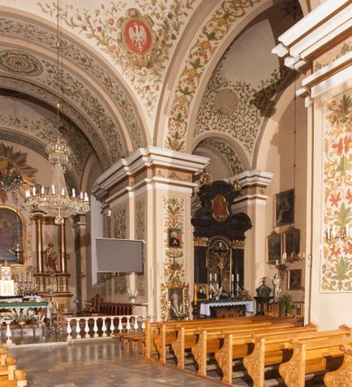 Zdjęcie nr 11: Kościół murowany, trójnawowy, halowy, na rzucie prostokąta z wydłużonym, skierowanym na północ prezbiterium z dobudowaną po zachodniej stronie zakrystią. Prezbiterium jednoprzęsłowe, węższe od korpusu, zamknięte prosto. Nawa główna oraz boczne dwuprzęsłowe, rozdzielone masywnymi opilastrowanymi filarami; artykulacja ścian przy pomocy filarów. Sklepienie prezbiterium kolebkowe, naw żagielkowe, przedzielone gurtami. Od południa chór muzyczny mieszczący się między pierwszymi filarami i powyżej kruchty wejściowej. W prezbiterium jedno okno od wschodu, drugie w zakrystii, w nawach bocznych po dwa. Główne wejście od południa flankowane przez dwa boczne, kolejne do zakrystii i między zakrystią i prezbiterium. Niektóre okna wypełnione gomułkami, ściany i sklepienia kościoła pokrywa polichromia utworzona z dekoracji floralnych i umieszczonych w jej obrębie symboli ewangelistów, herbów i putt wykonujących prace kojarzące się z regionem (bartnicze, ślusarskie, kowalskie itd.). Fasada dwuwieżowa, trójosiowa, dwuprzęsłowa, z portalem na każdej osi. Podziały pionowe przeprowadzono za pomocą półkolumienek i lizen, poziome w formie fryzu kostkowego wyniesionego nad środkowym portalem. Drzwi osi głównej w kształcie stojącego prostokąta ujętego półkolumnami dźwigającymi archiwoltę utworzoną z trzech łuków i trójkątny przyczółek. Drzwi osi bocznych zwieńczone przyczółkami. W drugiej kondygnacji na osi okulus i półokrągła konchowa wnęka z figurą Najświętszego Serca Jezusa poniżej trójkątnego szczytu dekorowanego fryzem arkadkowym. W osiach bocznych okna w kształcie stojącego prostokąta zamkniętego półkoliście. Wyżej dwie kondygnacje wież oddzielone gzymsem, w pierwszej  umieszczono okna, w drugiej tarcze zegarów. Elewacje boczne gładkie, przeprute jedynie oknami. Nawy przykrywa dach dwuspadowy, wieże namiotowe z fartuchem zwieńczone krzyżami.